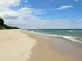 Plaża nad morzem, gdzie fale delikatnie obmywają piasek. W oddali widać zalesione wzgórze, a na niebie chmury. Idealne miejsce na spacer dla gąsek.