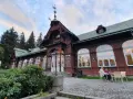 Drewniany pawilon z zegarem i karłowata studzienka przed wejściem. Na zewnątrz siedzą ludzie przy stoliku.